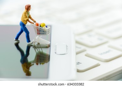 Miniature Shopper Pushes A Shopping Cart On A White Smart Phone. Concept Of Brick And Mortar Stores Nowadays Face With Increased Competition From Online Ecommerce And Decreasing In Foot Traffic.