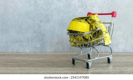 Miniature Shopping Cart with Yellow Measuring Tape on Wooden Surface Against Gray Background - Powered by Shutterstock