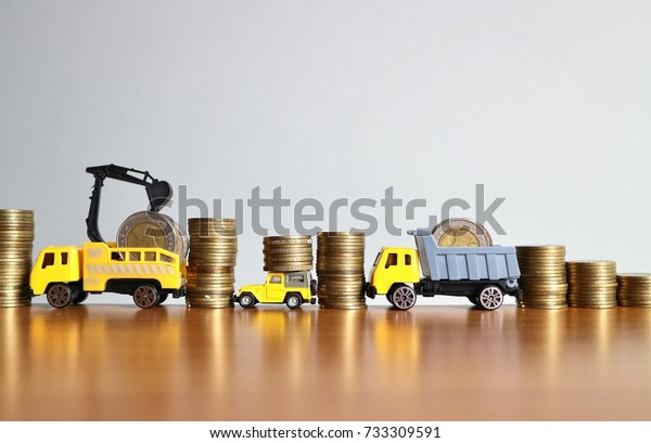 Miniature toy construction truck\
and car help carry rolls of gold coins money in line on wood table \
