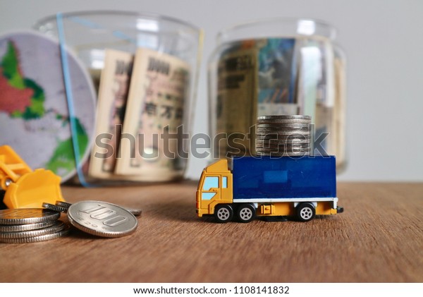 Miniature truck carry roll coins and Japan\
money on wood table with mixed pile of money inside bottle with\
world globe in blur grey\
background