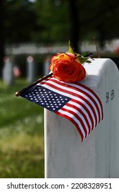 Miniature U.S. Flag And Pink Rose Resting On A Headstone In A Cemetary