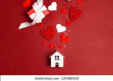 Miniature White Toy House With Hearts And Gifts On Red Background. St. Valentines Day Composition.