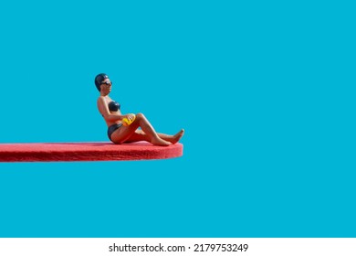 A Miniature Woman, Wearing A Black Bikingi, A Black Swimming Cap And Googles, Is Sitting On A Red Diving Board, On A Blue Background