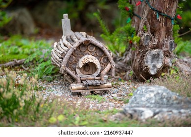 A Miniature Wooden Garden Gnome House In The Middle Of A Rich Green Garden. The House Has A Slanted Or Curved Root With A Peak Made From Shingles. The Walls Are Made From Small Cedar Shakes.