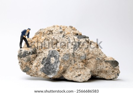 Miniature worker using a pickaxe to chip away at a large rock on white background