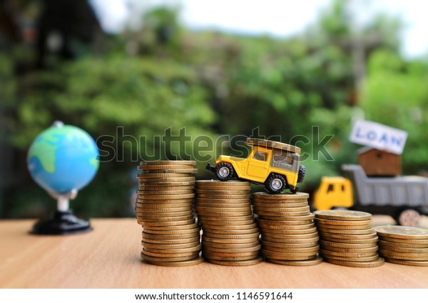 Miniature yellow car carry coin driving rolls of\
gold money and world globe with truck carry house with “loan” word\
on table in blur natural\
tree