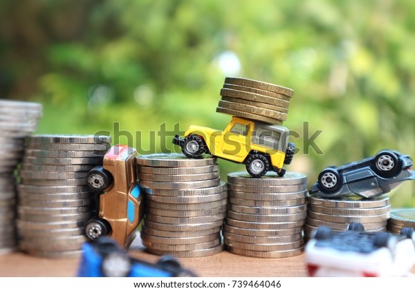 Miniature yellow car carry rolls\
coins and toy vehicles overturn upside down on rolls ladder of\
money on wood table in blur natural tree and bright light\
background