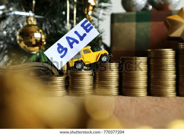 Miniature yellow car with “sale” on\
white paper drives  on rolls ladder of gold coin money in Christmas\
tree decorated with balls, silver ribbons on wood\
table