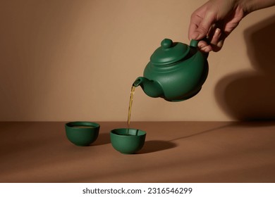 Minimal abstract background with set of teapot and cup. Against the brown background, female hand holding green pot and dropping tea in the cup. Front view, copy space - Powered by Shutterstock