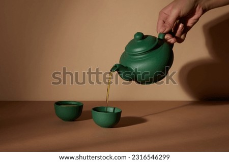 Minimal abstract background with set of teapot and cup. Against the brown background, female hand holding green pot and dropping tea in the cup. Front view, copy space