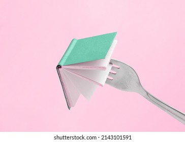 Minimal Abstract Composition Made Of Fork Holding A Green Book On Isolated Pastel Pink Background With Copy Space. Note Card For Book Fair, Promotions Or Education. Concept Of Reading Or Learning. 