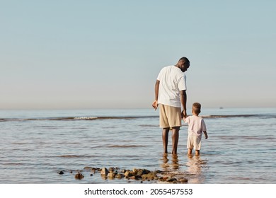 Minimal back view portrait of happy young father and son enjoying walk on beach together and standing in water, copy space