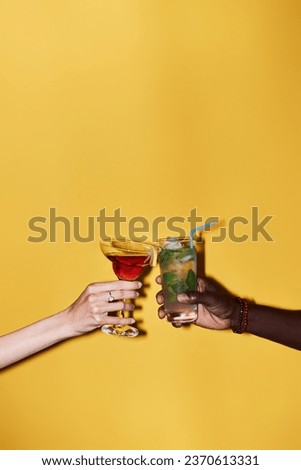 Minimal close up of two hands clinking cocktails against vibrant yellow background, copy space