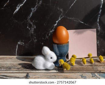 Minimal Easter still life featuring a brown egg in a blue ceramic cup, white bunny figurine, pussy willow branch and blank pink card on a rustic wooden surface. Clean spring composition with copy space and seasonal holiday charm.