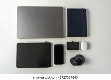 Minimal flat lay of modern tech essentials including laptop, tablet, smartphone, notebook, wallet, wireless earbuds, and headphones on a clean light background.