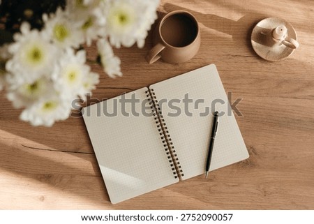 Minimal flat lay of an open grid notebook, coffee cup, and white chrysanthemum bouquet on a table, perfect for mindfulness, morning routine, work, study and cozy home lifestyle.