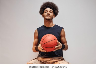 Minimal front view portrait of Black young man as basketball player holding ball and looking at camera sitting on bench against white wall copy space - Powered by Shutterstock