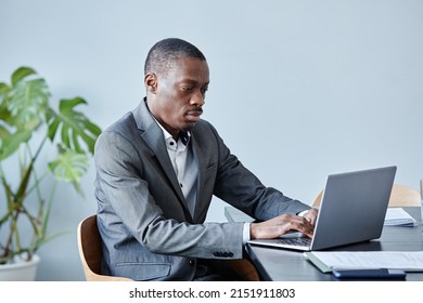 Minimal Portrait Of Professional Black Executive Using Laptop While Sitting At Workplace Against Blue Wall In Office And Wearing Suit, Copy Space