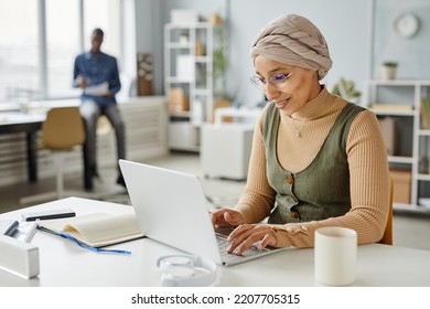 Minimal portrait of smiling Middle Eastern businesswoman wearing headwrap while typing at laptop in office, copy space - Powered by Shutterstock