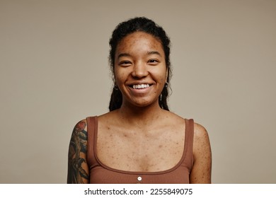 Minimal portrait of young black woman smiling at camera standing against neutral beige background with focus on real skin texture, spots and acne scars