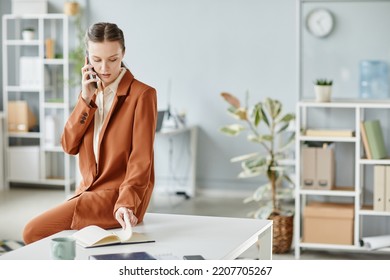 Minimal Portrait Of Young Female Boss Speaking By Smartphone While Sitting On Desk In White Office Interior, Copy Space