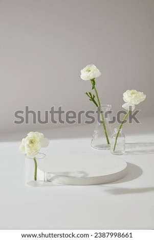 Minimal scene of white background featured few Persian Buttercup flowers inside glassware. Blank round podium to display your cosmetic product or goods