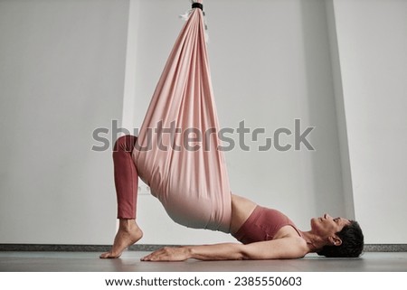 Minimal side view of mature woman enjoying aerial yoga and exercising with pink hammock, copy space