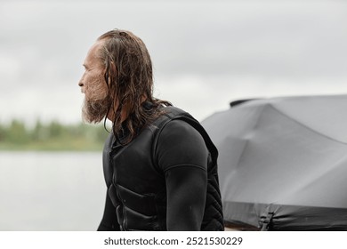 Minimal side view portrait of man wearing wetsuit and looking away sitting on back of boat in nature with long wet hair copy space