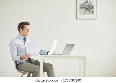 Minimal Side View Portrait Of Smiling Young Businessman Sitting At Desk And Working In Office Against White Wall, Copy Space