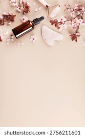 Minimal spa composition with amber essential oil bottle, jade roller, gua sha stone, bath salt and cherry blossom branches on beige background with copy space.