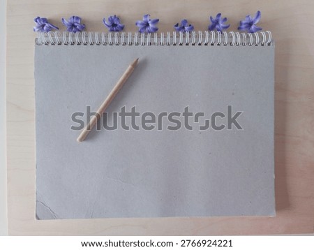Minimal spring composition on a light natural wooden background featuring a slightly worn spiral sketchbook with a gently creased corner, a natural cardboard pen placed on top, and a row of purple hyacinth blossoms arranged above the spiral. The scene represents creativity, new beginnings, fresh ideas and spring inspiration. The imperfect notebook adds authenticity, suggesting planning, sketching, brainstorming and creative process. Natural materials and soft lighting create a calm organic atmosphere suitable for seasonal themes, education, design workflow and lifestyle concepts. The composition communicates opportunity, personal growth, starting new projects and welcoming spring. Ideal for creative planning, education, stationery themes, blogging, marketing visuals and inspirational content. Clean layout with copy space supports editorial and advertising use. No people.