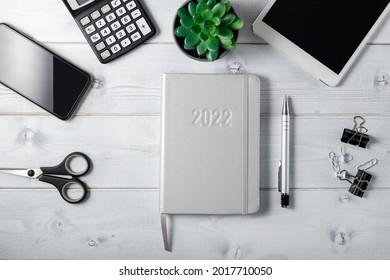 Minimal  unisex workplace with tablet,  silver notepad diary 2022, smartphone, small succulent and stationery on a light whie grey  wooden background. At home or in office. Happy New 2022 Year card