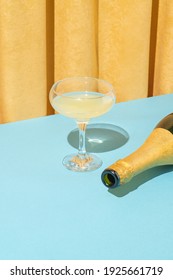 Minimal Vintage Scene With Champagne Glass And Bottle On Pastel Blue Table Against Yellow Velvet Curtain. Bright Light, Retro Party.