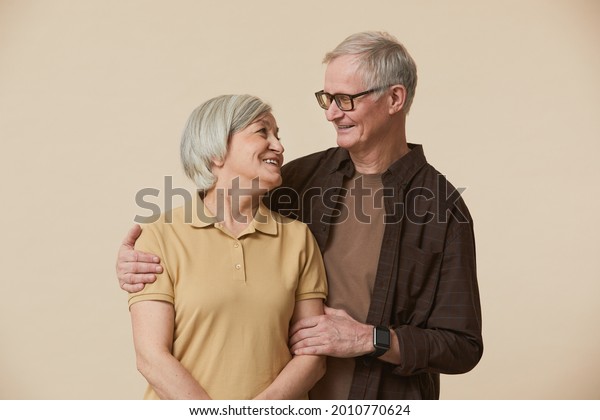 Minimal waist up portrait of carefree senior\
couple embracing and looking at each other while standing against\
beige background