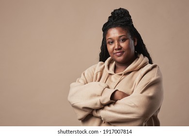 Minimal Waist Up Portrait Of Confident Curvy Woman Looking At Camera And Wearing Comfortable Hoodie While Standing With Arms Crossed Against Neutral Beige Background In Studio, Copy Space