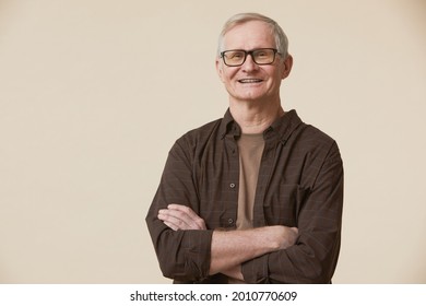 Minimal waist up portrait of senior man wearing glasses and looking at camera while standing with arms crossed against beige background - Powered by Shutterstock
