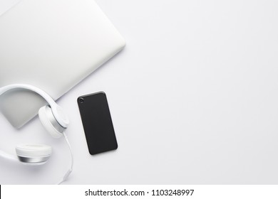 Minimal Work Space Concept. Top View Of Laptop, White Headphones And Black Cell Phone On White Background With Copy Space. Flat Lay.