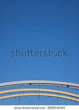 Minimalist architectural composition featuring the curved steel arches of a modern bridge at the bottom of the frame. The vast majority of the image is filled with a solid, vibrant clear blue sky, providing ample negative space (copy space) for text and graphic design