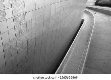 A minimalist black and white architectural photo showing a curved wall with textured panels, leading to a distant solitary figure walking along a curved path. - Powered by Shutterstock