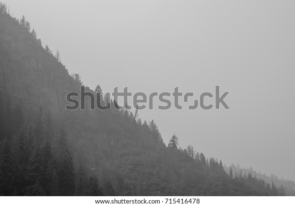 Minimalist\
black and white photo of the side of a mountain dividing diagonally\
the picture to a dark and a bright side. Silhouettes of pine trees\
are visible on the edge. Captured in\
Yosemite.