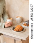 A minimalist coffee scene with two cappuccinos in ribbed ceramic mugs and two pastries: a golden croissant and a chocolate croissant. A person in a green sweater holds one cup, adding a cozy touch.