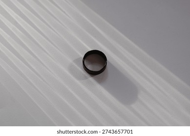 Minimalist composition of a black ring placed on a textured white surface with soft natural window light creating elegant shadow lines.