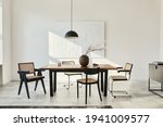 Minimalist composition of dining room interior with wooden table, design chairs, dried flowers in a vase, black pendant lamp, art paintings on the wall and elegant personal accessories. Template.