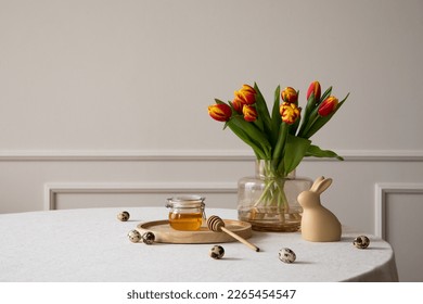 Minimalist composition of easter dining room interior with copy space, round table, vase with tulips, easter bunny sculpture, honey in jar, beige wall and personal accessories. Home decor. Template.
