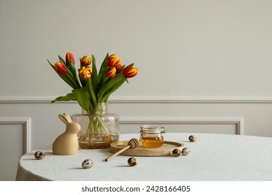 Minimalist composition of easter dining room interior with copy space, round table, vase with tulips, easter bunny sculpture, honey in jar, beige wall and personal accessories. Home decor. Template.