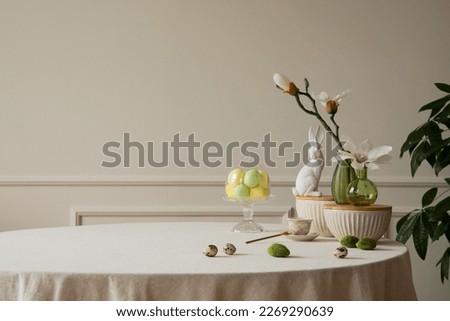 Minimalist composition of easter dining room interior with copy space, gray easter bunny sculpture, colorful eggs, vase with magnolia, beige wall with stucco and accessories. Home decor. Template.