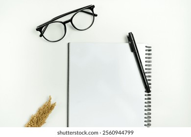 A minimalist composition featuring a blank page of a spiral-bound notebook accompanied by a pen, glasses, and dried plants, all placed on a plain white surface