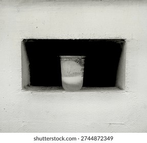 The minimalist composition featuring a transparent plastic cup placed inside a rectangular recess in a white concrete wall. The cup contains a small amount of white liquid at the bottom, with condensation and residue visible along its inner surface. A thin plastic seal remains partially attached to the rim.

The recessed space behind the cup is dark, creating a strong contrast with the bright white wall and drawing attention to the solitary object. The rough texture of the concrete surface adds subtle detail and depth to the otherwise simple setting.
