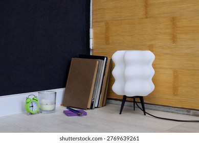 A minimalist desk setup featuring a unique wavy lamp, a stack of books, a candle, and a bright green alarm clock