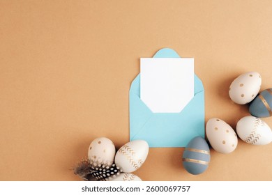 Minimalist Easter composition with a blue envelope, blank white card, and decorative eggs with golden patterns on a neutral brown background. Top view, flat lay, mockup.